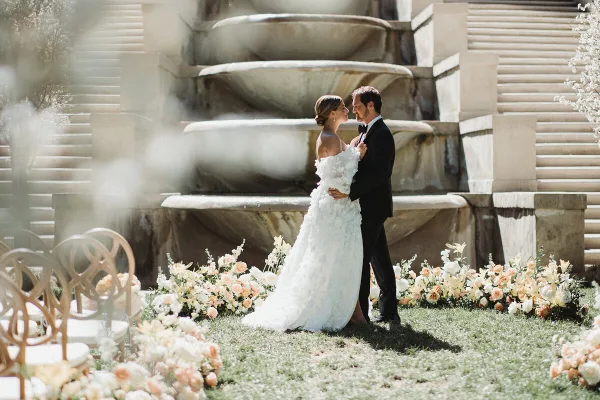 A bride and groom embracing in front of a stone fountain and floral arrangements during a wedding.