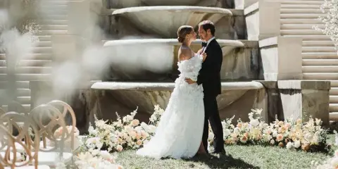 A bride and groom embracing in front of a stone fountain and floral arrangements during a wedding.