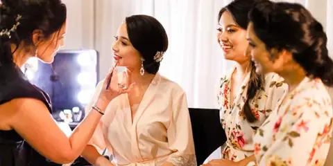 A makeup artist applying makeup to a bride while bridesmaids look on in a bridal suite.