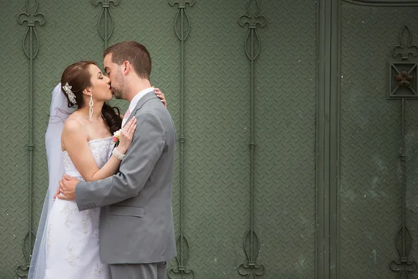 Bride in white gown and groom in gray suit kissing in front of a green textured wall.
