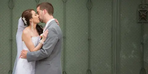 Bride in white gown and groom in gray suit kissing in front of a green textured wall.