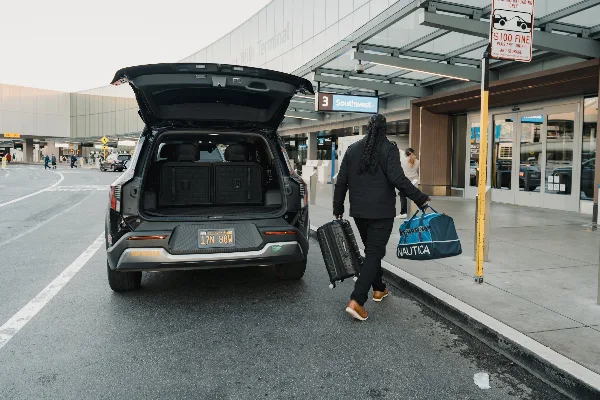 A driver carrying luggage toward a Kia EV9 SUV with open trunk at an airport terminal curb.