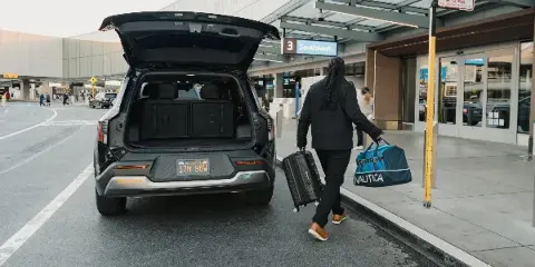 A driver carrying luggage toward a Kia EV9 SUV with open trunk at an airport terminal curb.