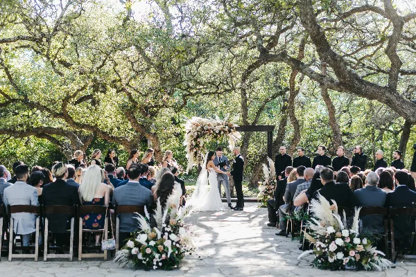 A wedding ceremony taking place outdoors under large oak trees, featuring a floral arch, seated guests, and the couple exchanging vows.