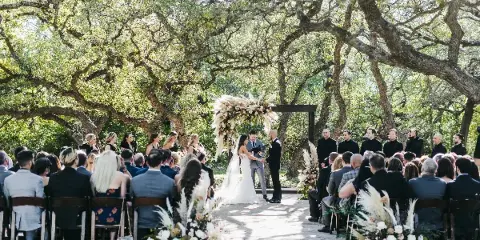 A wedding ceremony taking place outdoors under large oak trees, featuring a floral arch, seated guests, and the couple exchanging vows.