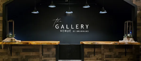 Interior view of The Gallery Venue by Branhams showing a dark feature wall with the venue name, pendant lighting, and rustic wood counter surfaces.