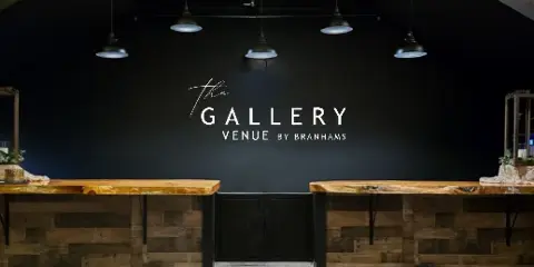 Interior view of The Gallery Venue by Branhams showing a dark feature wall with the venue name, pendant lighting, and rustic wood counter surfaces.