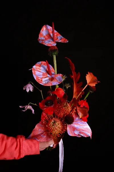 A hand holding a modern, sculptural floral arrangement with red and pink patterned elements against a black background.