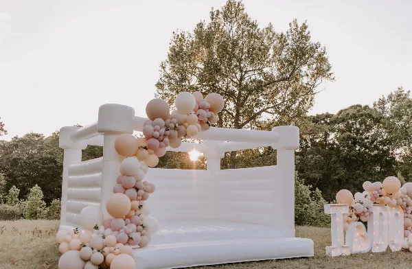 A white luxury bounce house set up in a grassy field, decorated with a pink and white balloon garland and a light-up sign.
