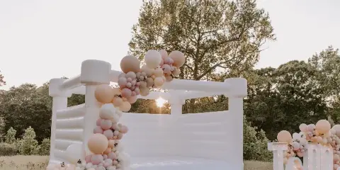 A white luxury bounce house set up in a grassy field, decorated with a pink and white balloon garland and a light-up sign.