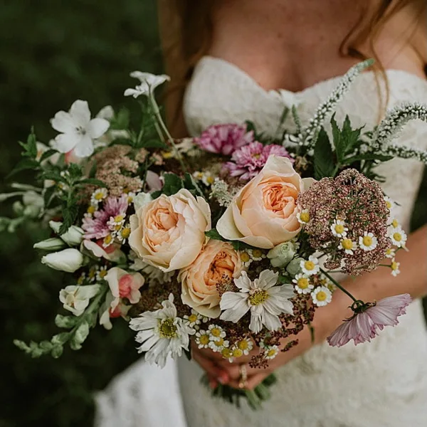 A bride in a white wedding dress holding a large, rustic bouquet of cream roses, daisies, and greenery.