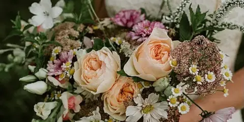 A bride in a white wedding dress holding a large, rustic bouquet of cream roses, daisies, and greenery.