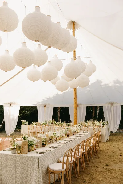 A wedding tent interior decorated with hanging white paper lanterns, white fabric draping, and long banquet tables with floral centerpieces.