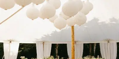 A wedding tent interior decorated with hanging white paper lanterns, white fabric draping, and long banquet tables with floral centerpieces.
