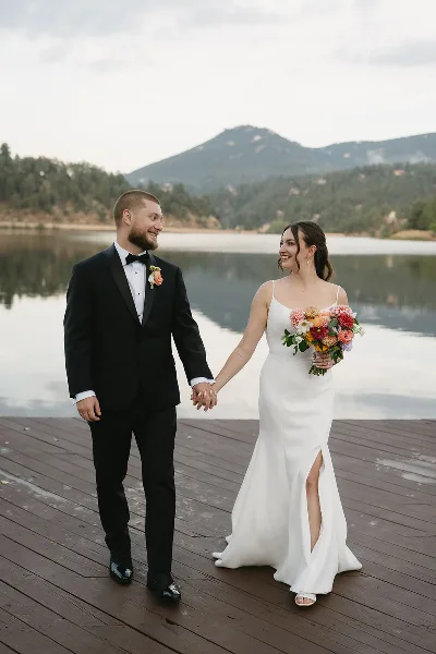 A bride and groom holding hands and smiling at each other on a wooden dock with a lake and mountains in the background.
