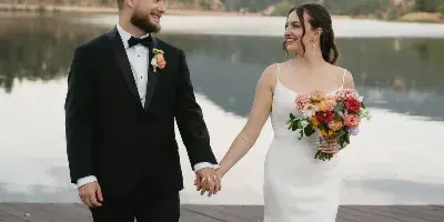 A bride and groom holding hands and smiling at each other on a wooden dock with a lake and mountains in the background.