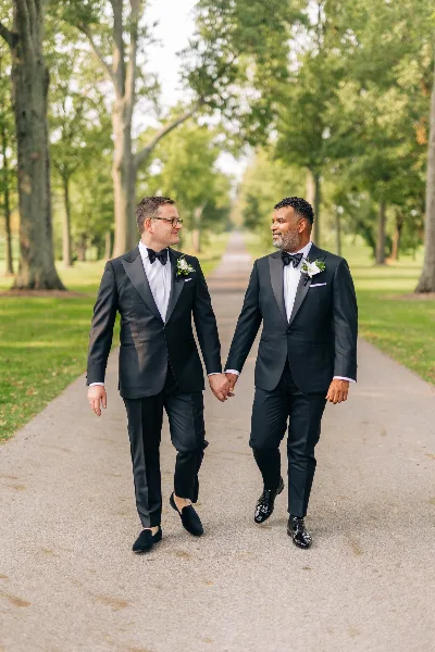 Two men in black tuxedos walking hand-in-hand on a tree-lined path.