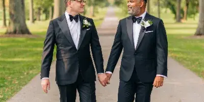 Two men in black tuxedos walking hand-in-hand on a tree-lined path.