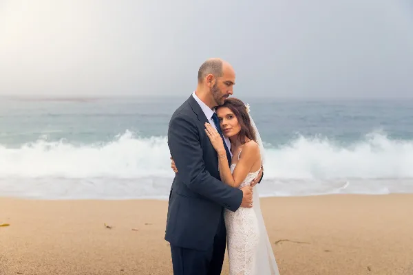 A bride and groom embracing on a sandy beach with ocean waves in the background.