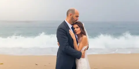 A bride and groom embracing on a sandy beach with ocean waves in the background.