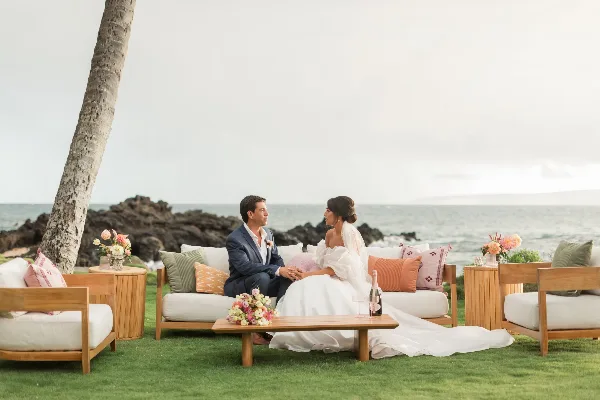 A bride and groom sitting on a wooden outdoor lounge set with white cushions and colorful pillows, set against a beach backdrop.