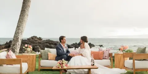 A bride and groom sitting on a wooden outdoor lounge set with white cushions and colorful pillows, set against a beach backdrop.