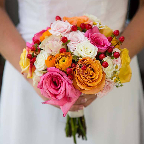 A close-up of a colorful wedding bouquet featuring pink, orange, and white roses and ranunculus, held by a bride in a white dress.