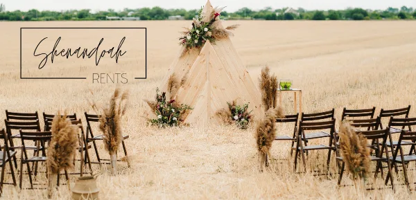 Outdoor wedding ceremony setup with a wooden teepee, floral arrangements, and rows of wooden chairs in a field.