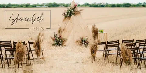 Outdoor wedding ceremony setup with a wooden teepee, floral arrangements, and rows of wooden chairs in a field.