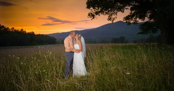 A bride and groom embracing in a grassy field with a mountain range and sunset in the background.