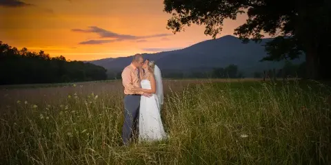 A bride and groom embracing in a grassy field with a mountain range and sunset in the background.