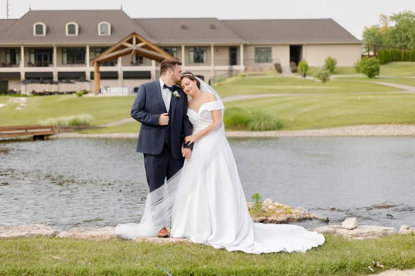 A bride and groom standing together outdoors near a pond with a large venue building in the background.