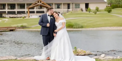 A bride and groom standing together outdoors near a pond with a large venue building in the background.