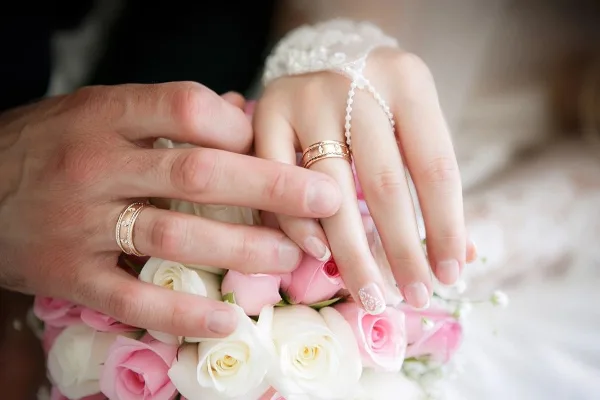Close-up of a couple's hands exchanging wedding rings over a pink and white rose bouquet.