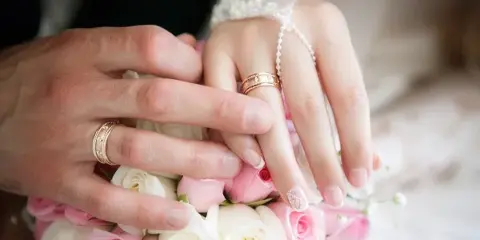 Close-up of a couple's hands exchanging wedding rings over a pink and white rose bouquet.
