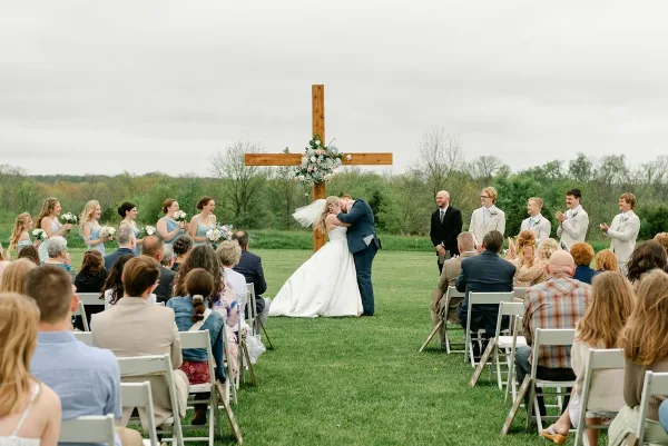 A bride and groom sharing a kiss at an outdoor wedding ceremony in a grassy field with a wooden cross and guests seated in white chairs.
