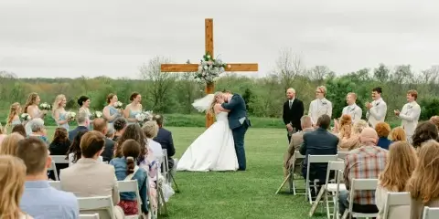 A bride and groom sharing a kiss at an outdoor wedding ceremony in a grassy field with a wooden cross and guests seated in white chairs.