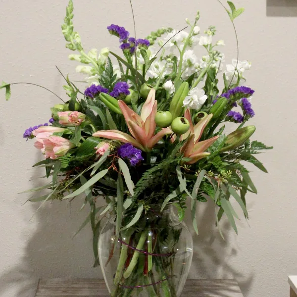 A tall floral arrangement with pink lilies, purple stock, and white flowers in a clear glass vase.
