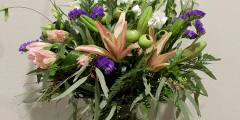 A tall floral arrangement with pink lilies, purple stock, and white flowers in a clear glass vase.
