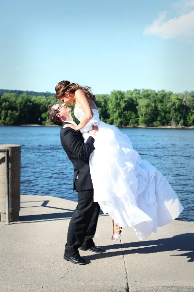 A groom in a black suit lifts a bride in a white wedding dress in a dip pose on a concrete pier with a lake and trees in the background.