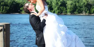 A groom in a black suit lifts a bride in a white wedding dress in a dip pose on a concrete pier with a lake and trees in the background.