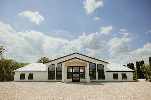 Exterior view of a modern white wedding venue building with a metal roof and large windows, surrounded by trees and a gravel parking area.