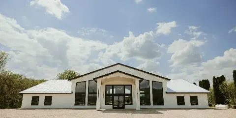 Exterior view of a modern white wedding venue building with a metal roof and large windows, surrounded by trees and a gravel parking area.