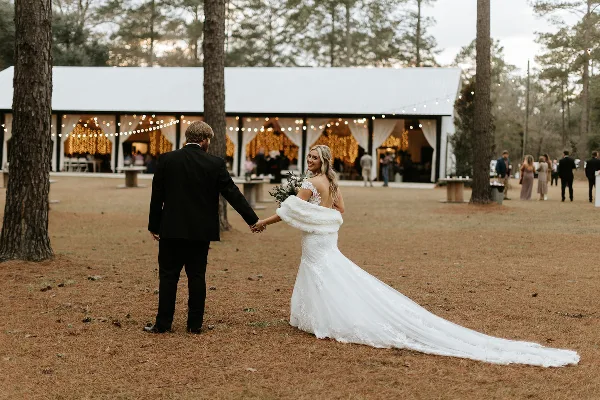 A bride and groom holding hands in a pine forest with a white barn reception venue in the background.