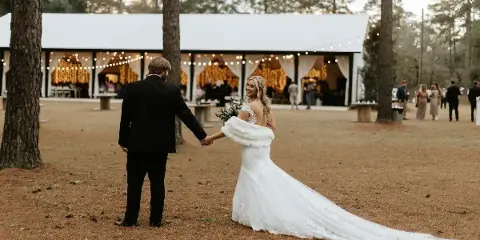 A bride and groom holding hands in a pine forest with a white barn reception venue in the background.