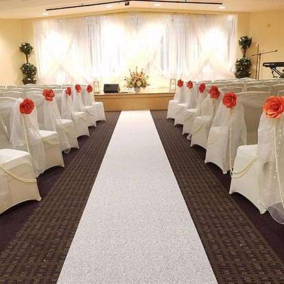 A decorated wedding aisle with white chair covers and floral arrangements leading to a stage with a white backdrop.