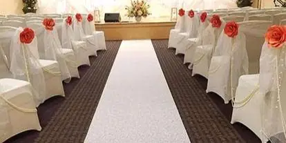 A decorated wedding aisle with white chair covers and floral arrangements leading to a stage with a white backdrop.