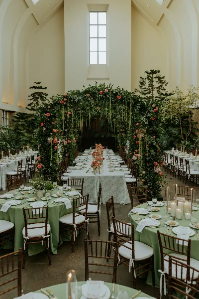 Styled wedding reception with long tables, green linens, wooden chairs, candles, and a lush floral arch backdrop.