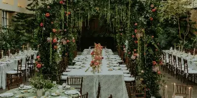 Styled wedding reception with long tables, green linens, wooden chairs, candles, and a lush floral arch backdrop.