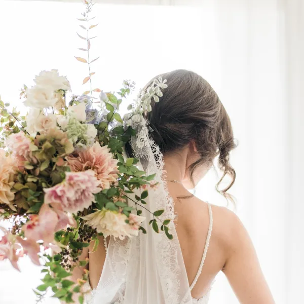 A bride from behind holding a large, romantic bouquet of pink and white flowers with greenery, wearing a floral hair accessory.